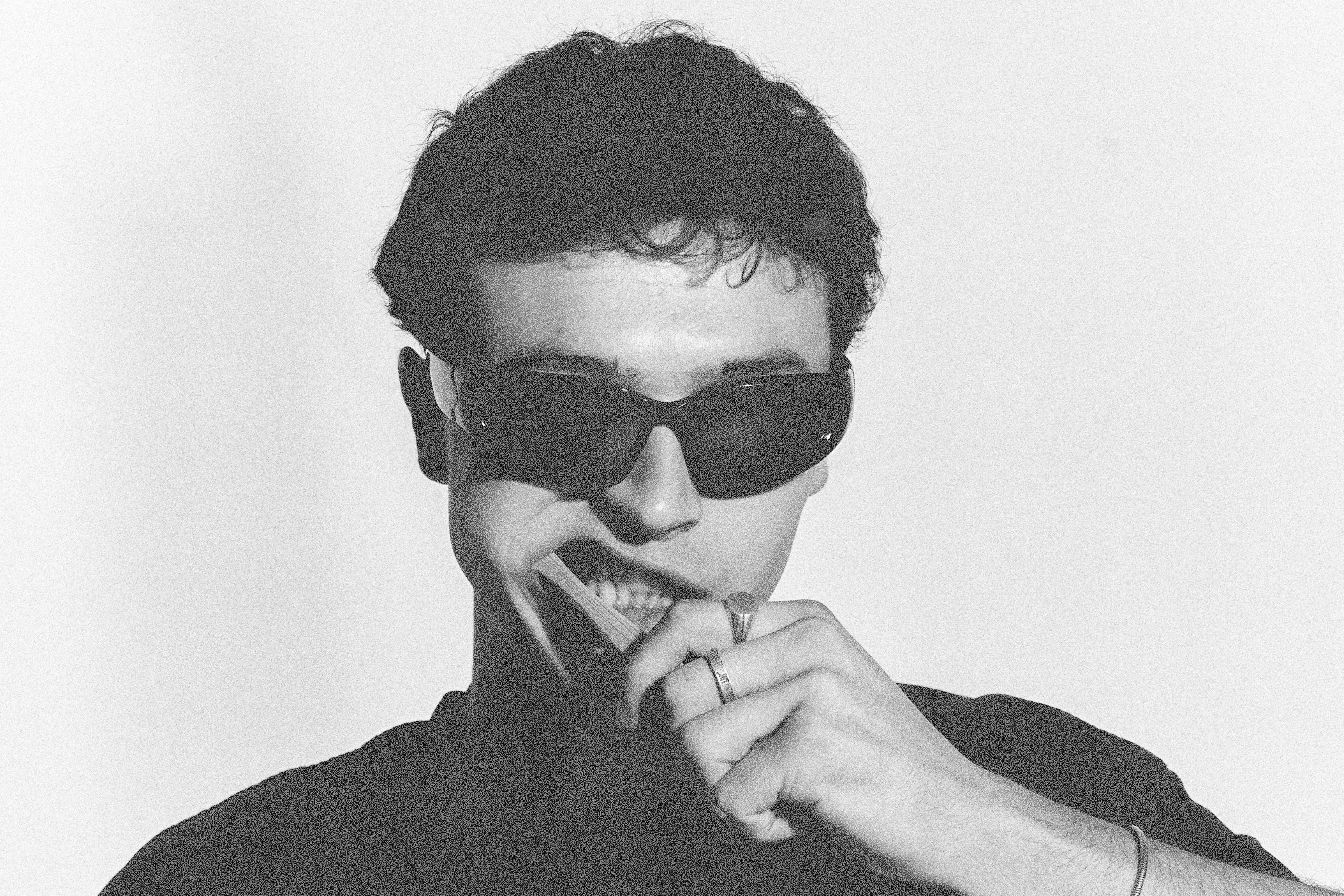 Black and white photo of a person wearing sunglasses and a dark shirt, and blue serpent rings and necklace and jewelry 