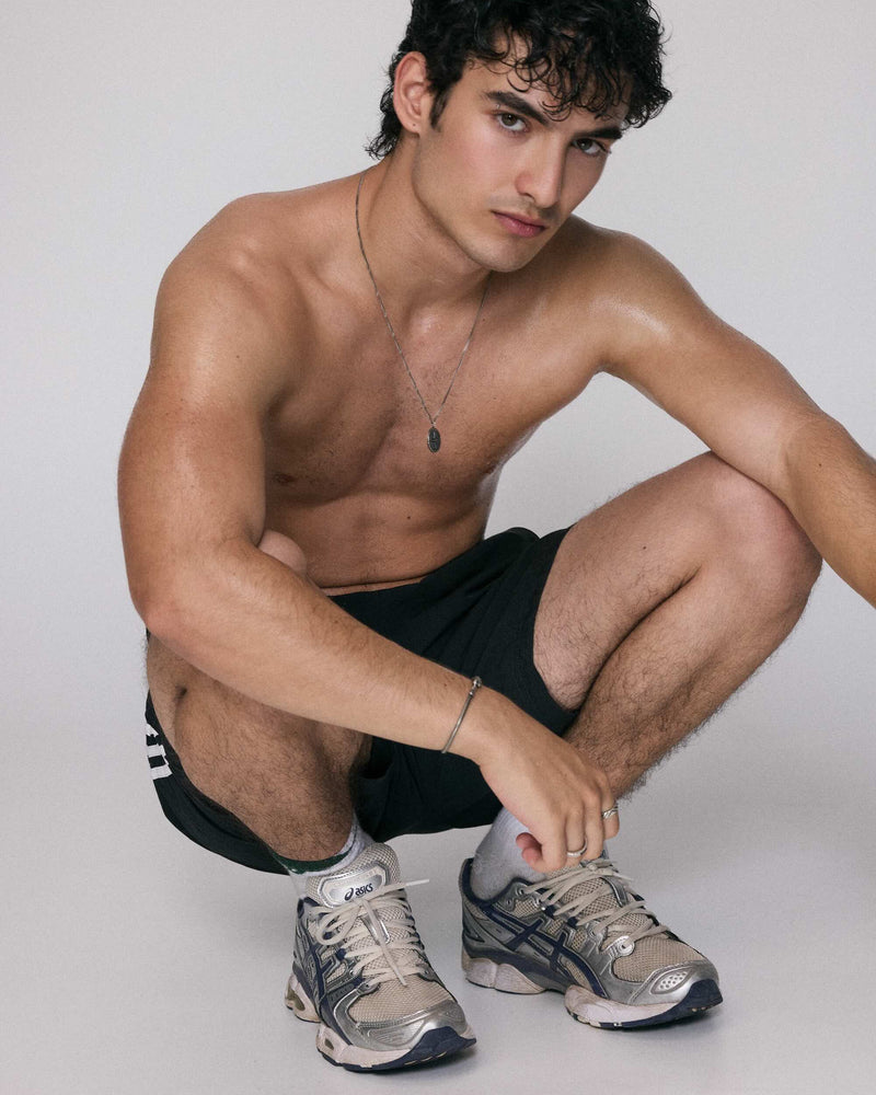Man sitting on a white floor wearing black shorts and gray sneakers, posing against a plain background wearing 925 silver necklace nodes