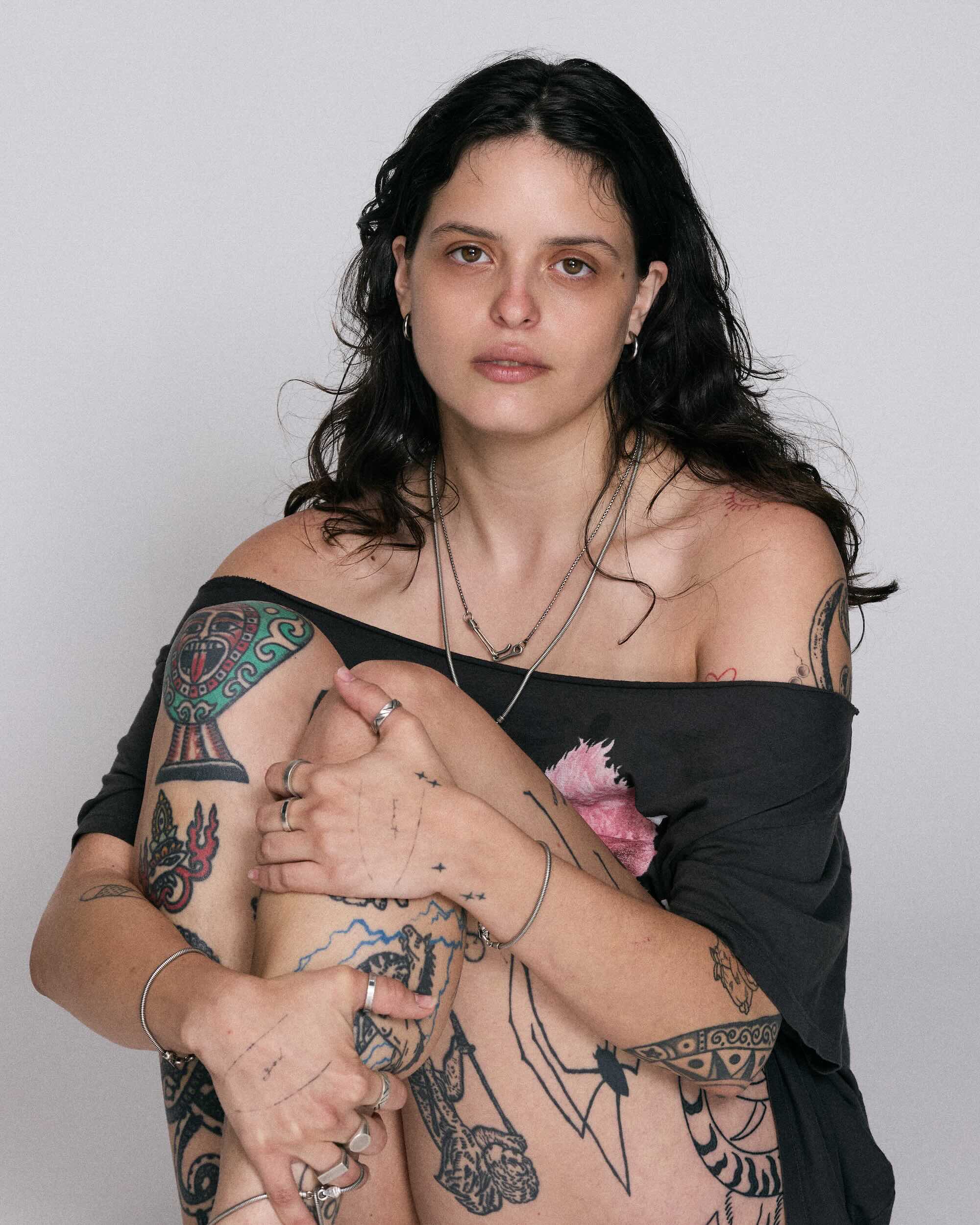 Woman with tattoos on her arms and chest against a plain background wearing 925 blue serpent rings and necklaces