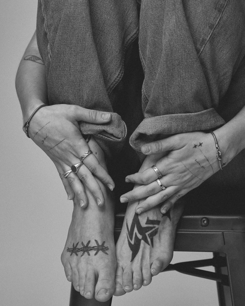Black and white photo of a person sitting with tattoos on their hands and feet wearing 925 blue serpent jewelry